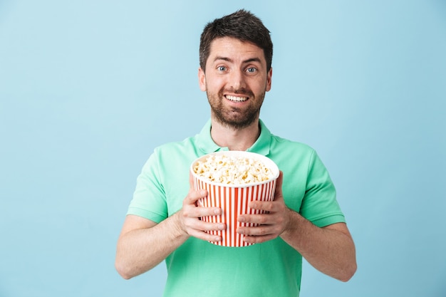 Портрет красивого бородатого мужчины в повседневной одежде, стоящего изолированно над синей стеной и поедающего попкорн во время просмотра фильма