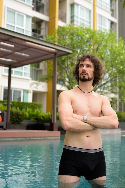 Portrait of handsome bearded man with curly hair shirtless relaxing beside the swimming pool in the city of Bangkok, Thailand