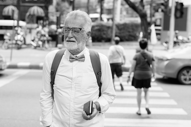 Portrait of handsome bearded senior tourist man wearing stylish clothes while exploring the city of Bangkok in black and white