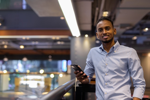 Portrait of handsome black African businessman inside shopping mall smiling and holding mobile phone horizontal shot