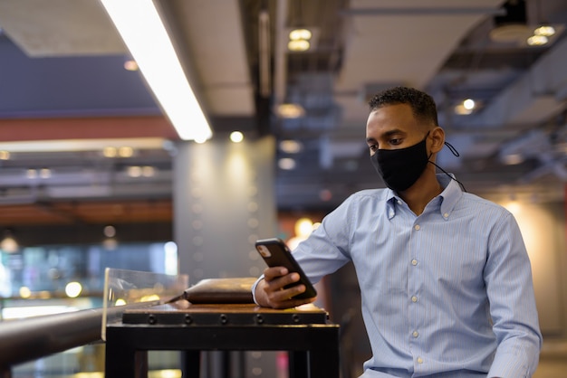 Portrait of handsome black African businessman inside shopping mall wearing face mask and using mobile phone horizontal shot