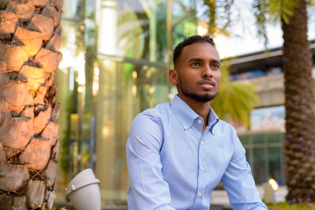 Portrait of handsome black African businessman outdoors in city during summer sitting and thinking horizontal shot