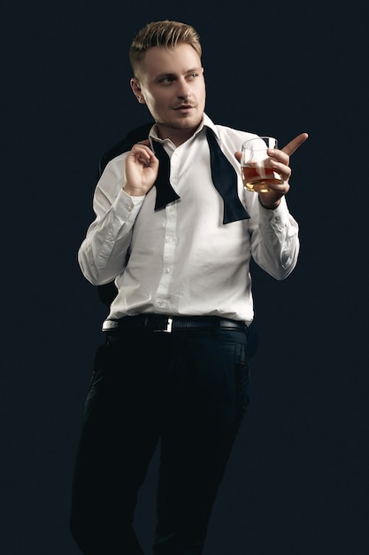 Portrait of handsome blond man in elegant tuxedo and bow tie posing with glass of whiskey on black  wall