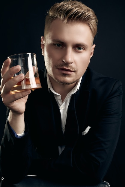 Photo portrait of handsome blond man in elegant tuxedo and bow tie posing with glass of whiskey on black  wall