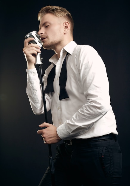 Ritratto del cantante uomo biondo bello in smoking elegante e farfallino in posa con microfono vintage sulla parete nera