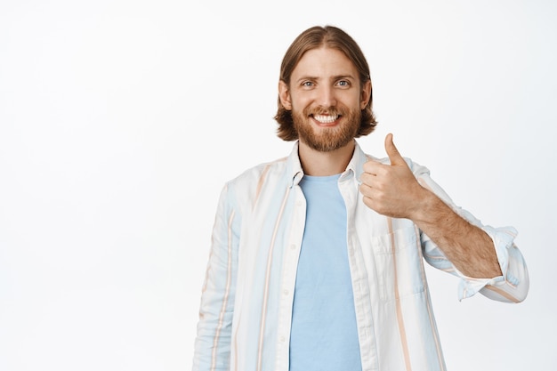 Portrait of handsome blond man smiling, showing thumb up with confident happy face, approve and agree, like something good, stanidng on white