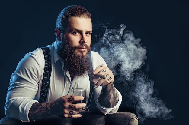 Портрет красивого брутального бородатого мужчины с сигарой и стаканом виски в темной комнате татуирует руки