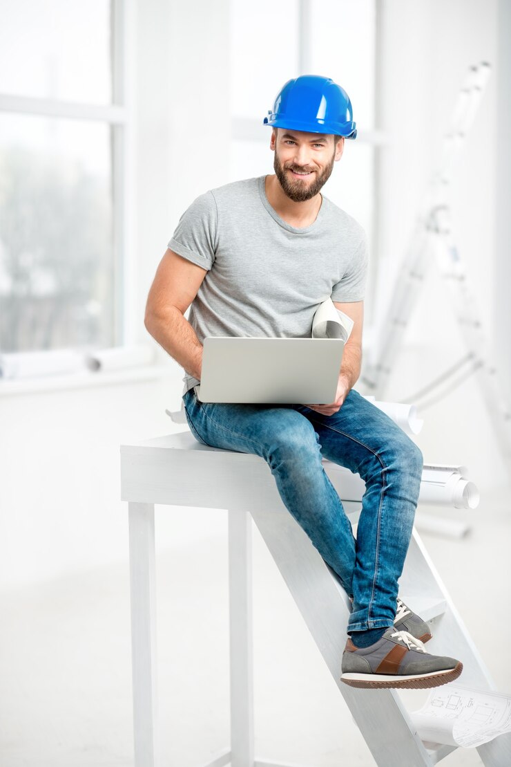 Portrait of a handsome builder, foreman or repairman in the helmet sitting with laptop in the white interior