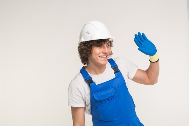 Portrait of handsome builder in white helmet and blue overall shaking his hand and laughing on white wall with copy space