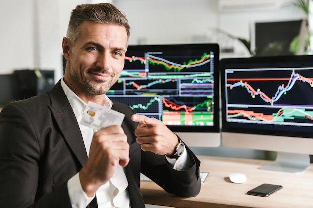 Portrait of handsome businessman 30s wearing suit holding credit card while sitting in office and working with digital graphics on computer