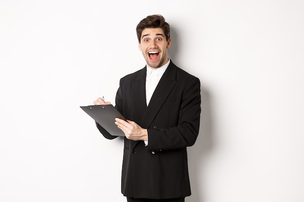 Portrait of handsome businessman in black suit, looking amazed while signing documents on clipboard, standing against white background.