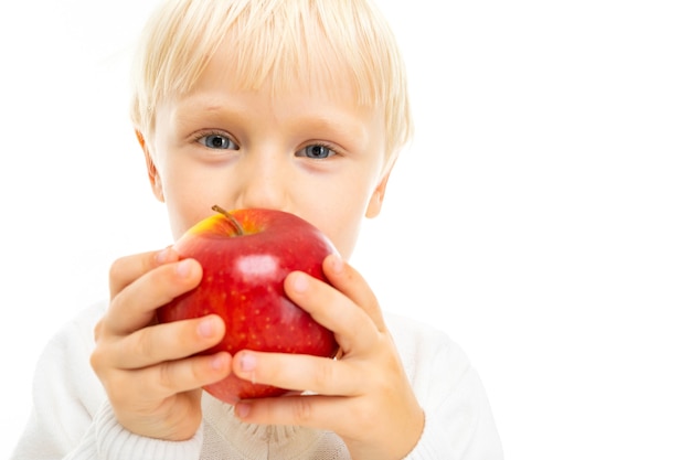 Portrait of handsome caucasian boy with blonde hair and white skin in white blouse and blue shorts eats a red apple