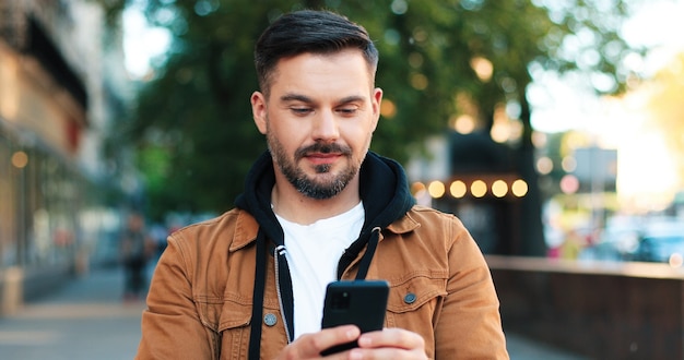 Portrait of the handsome caucasian man showing charming smile\
and healthy teeth while walking with his smartphone at the street.\
people and gadgets concept