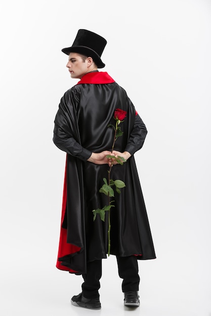 Photo portrait of handsome caucasian vampire holding red beautiful rose on white studio background.