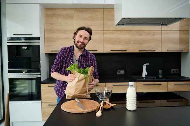 Portrait of a handsome charming Caucasian man with eco papr bag full of healthy food, greens and whole grain bread standing by a kitchen table in his spacious smart villa