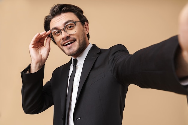 Portrait of a handsome cheerful young businessman wearing suit standing isolated over beige wall, taking a selfie