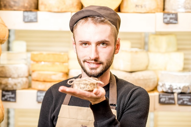 Portrait of a handsome cheese seller in uniform holding young goat cheese in front of the store showcase full of goods