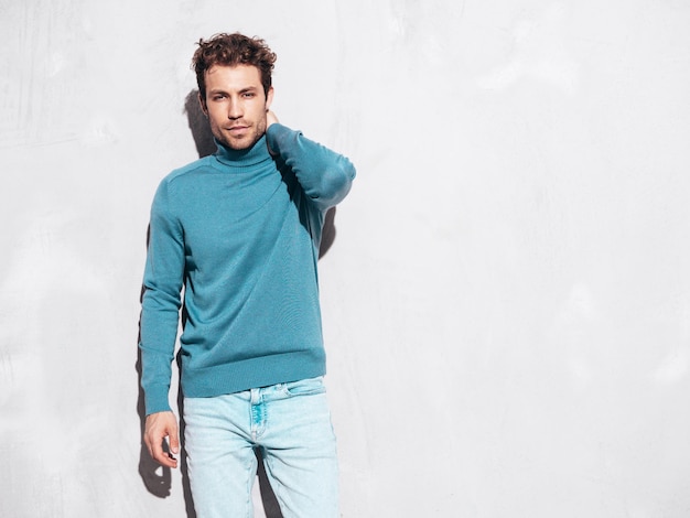 Portrait of handsome confident model Sexy stylish man dressed in blue sweater and jeans Fashion hipster male with curly hairstyle posing near grey wall in studio Isolated