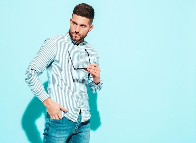 Portrait of handsome confident model Sexy stylish man dressed in shirt and jeans Fashion hipster male posing near blue wall in studio