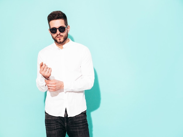 Portrait of handsome confident model Sexy stylish man dressed in white shirt and jeans Fashion hipster male posing near blue wall in studio In sunglasses Isolated