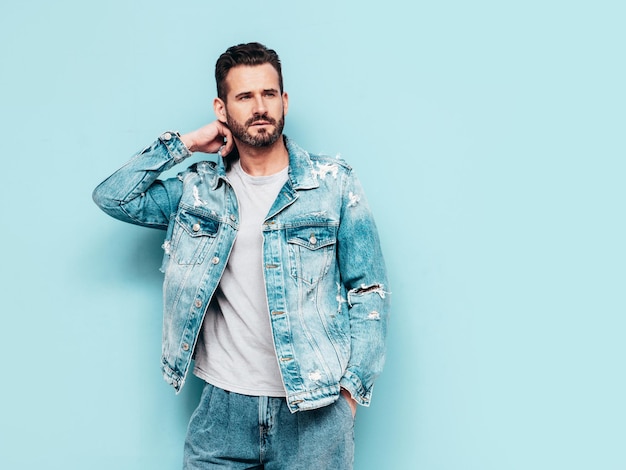 Portrait of handsome confident stylish hipster lambersexual model Sexy man dressed in jeans jacket Fashion male isolated on blue wall in studio