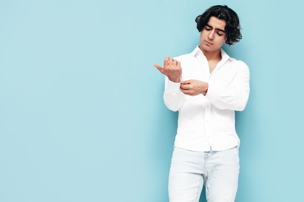 Portrait of handsome confident stylish hipster lambersexual model Sexy man dressed in summer white shirt and jeans clothes Fashion male isolated in studio Posing near blue wall