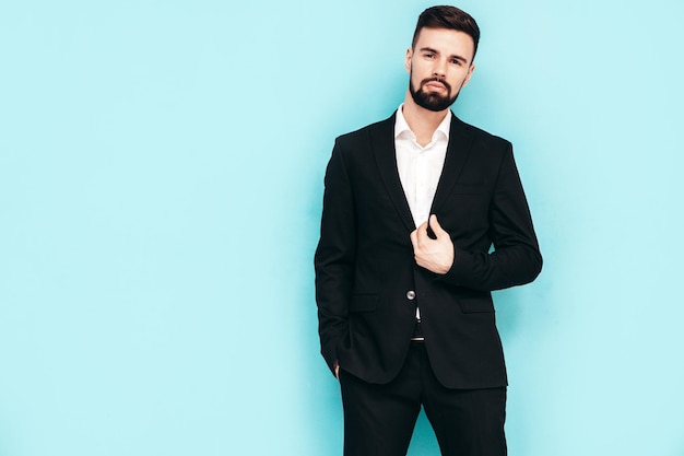 Portrait of handsome confident stylish hipster lambersexual model sexy modern man dressed in elegant black suit fashion male posing in studio near blue wall