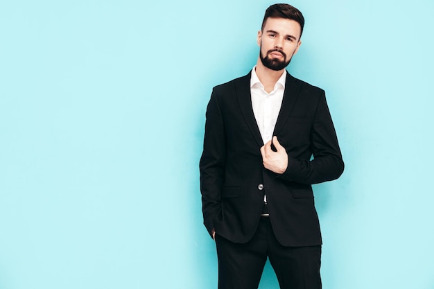 Portrait of handsome confident stylish hipster lambersexual model Sexy modern man dressed in elegant black suit Fashion male posing in studio near blue wall