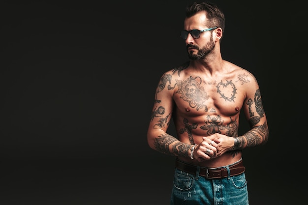 Portrait of handsome confident stylish hipster lambersexual model Sexy modern man Naked torso with tattoosFashion male posing in studio on dark background In sunglasses