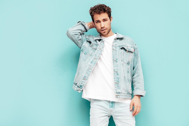 Portrait of handsome confident stylish hipster lambersexual model with curly hairstyle Sexy man dressed in jeans jacket Fashion male isolated on blue wall in studio