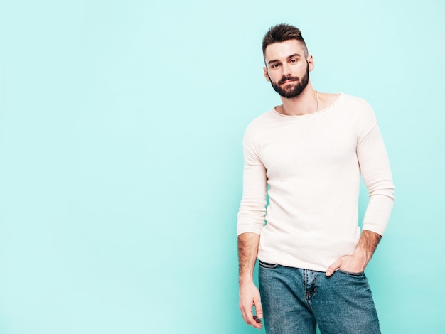 Portrait of handsome confident stylish hipster lambersexual modelMan dressed in white sweater and jeans Fashion male isolated on blue wall in studio Thoughtful