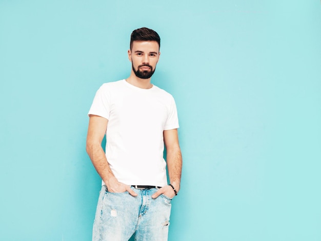 Portrait of handsome confident stylish hipster lambersexual modelMan dressed in white Tshirt and jeans Fashion male isolated on blue wall in studio