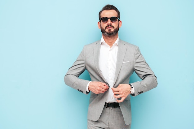 Portrait of handsome confident stylish hipster lambersexual modelModern man dressed in elegant suit Fashion male posing in studio near blue wall in sunglasses