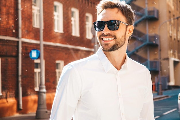 Portrait of handsome confident stylish hipster lambersexual modelModern man dressed in white shirt Fashion male posing on the street background in sunglasses Outdoors at sunset