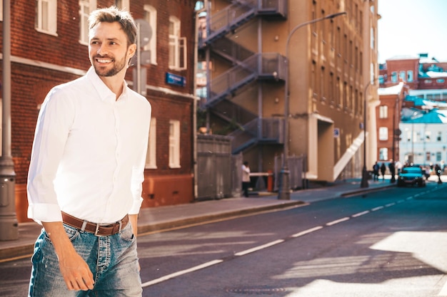 Portrait of handsome confident stylish hipster lambersexual modelModern man dressed in white shirt Fashion male posing on the street background in sunglasses Outdoors at sunset