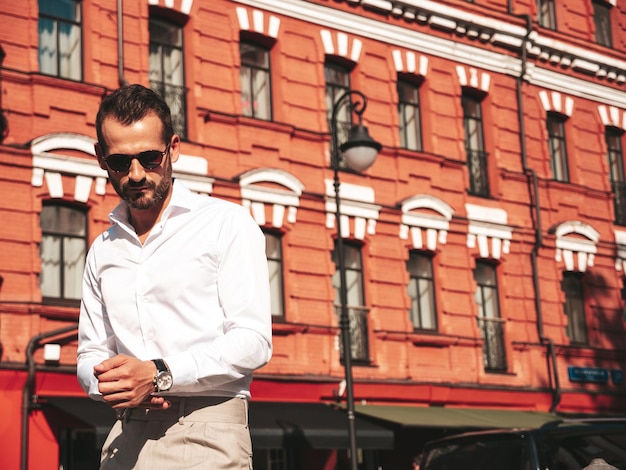 Portrait of handsome confident stylish hipster lambersexual modelModern man dressed in white shirt and trousers Fashion male posing in the street background in Europe city at sunset In sunglasses