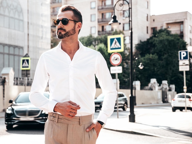 Portrait of handsome confident stylish hipster lambersexual modelModern man dressed in white shirt and trousers Fashion male posing in the street background in Europe city at sunset In sunglasses