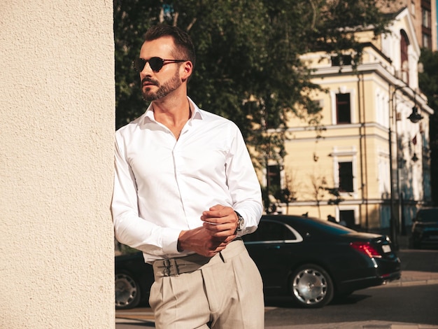 Portrait of handsome confident stylish hipster lambersexual modelModern man dressed in white shirt and trousers Fashion male posing in the street background in Europe city at sunset In sunglasses