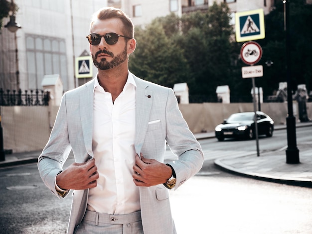 Portrait of handsome confident stylish hipster lambersexual modelSexy modern man dressed in elegant white suit Fashion male posing in the street background in Europe city at sunset In sunglasses