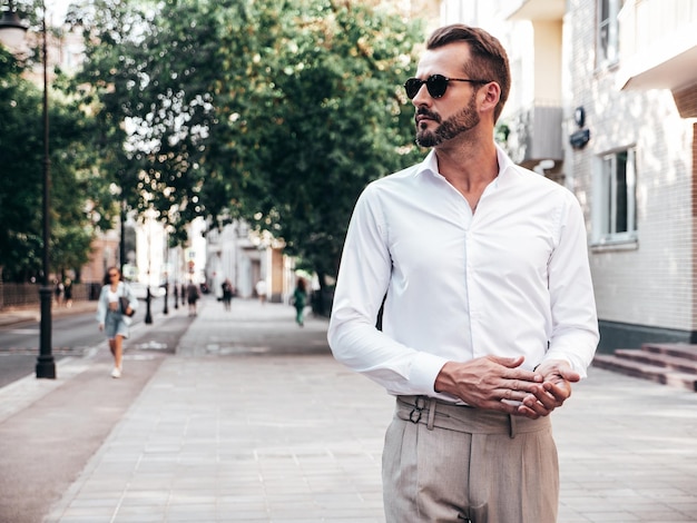 Portrait of handsome confident stylish hipster lambersexual modelSexy modern man dressed in white shirt and trousers Fashion male posing on street background in Europe city at sunset In sunglasses