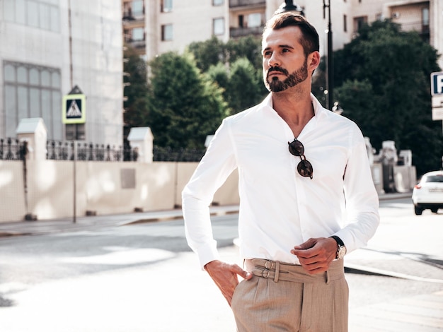 Portrait of handsome confident stylish hipster lambersexual modelSexy modern man dressed in white shirt and trousers Fashion male posing on street background in Europe city at sunset In sunglasses