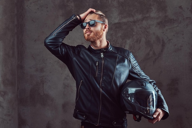 Portrait of a handsome confident stylish redhead biker in a black leather jacket and sunglasses, holds motorcycle helmet, posing in a studio. Isolated on a dark background.