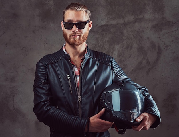 Portrait of a handsome confident stylish redhead biker in a black leather jacket and sunglasses, holds motorcycle helmet, posing in a studio. Isolated on a dark background.