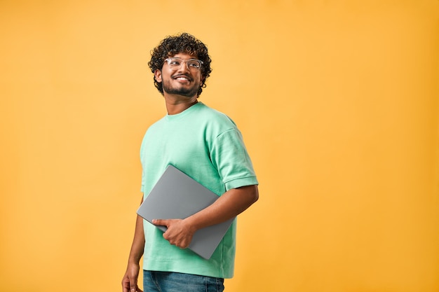 Портрет красивого кудрявого индийского мужчины в бирюзовой футболке и очках, смеющегося, отводящего взгляд и держащего в руках ноутбук. Копирование пространства
