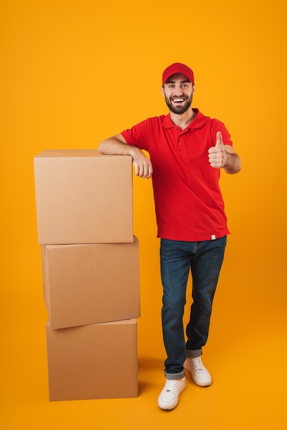 Portrait of handsome delivery man in red uniform showing thumb up while standing with packaging boxes isolated over yellow