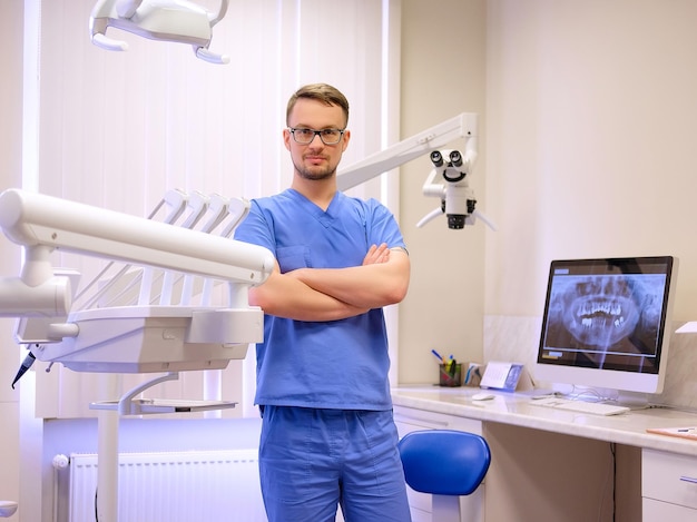 Portrait of a handsome dentist wearing a blue uniform, standing in a dentist clinic. Looking at camera.