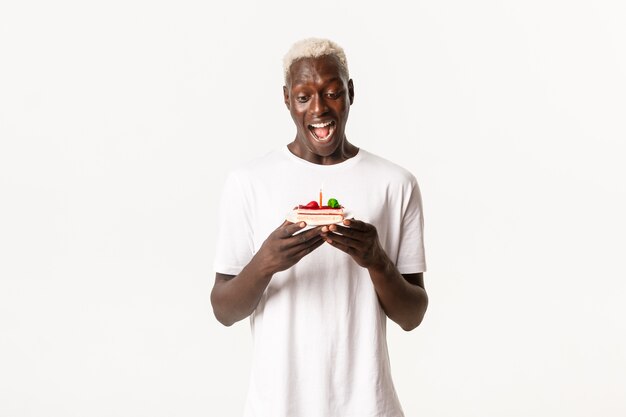 Portrait of handsome, excited african-american blond man, looking amused at birthday cake, smiling and making wish on lit candle