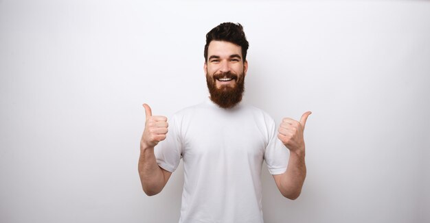 Portrait of handsome and excited bearded man showing thumbs up over white wall