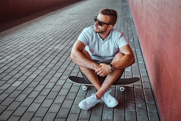 Portrait of a handsome fashionable skater guy in sunglasses dressed in a white shirt and shorts sitting on a skateboard under the bridge, looking away.