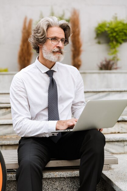 Portrait of handsome focused businessman in eyeglasses typing on laptop while sitting on bench outdoors
